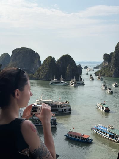 The image captures a scenic view of Ha Long Bay, Vietnam, on a clear day, with a person actively engaged in photography in the foreground. The perspective is from an elevated vantage point looking out over the water and islands.
In the foreground, viewed from the right profile, is a person with dark hair, wearing sunglasses pushed up onto their head and a dark-colored top. They are holding a small, silver digital camera with both hands, aiming it towards the picturesque landscape. Visible on their right arm are several tattoos, including a large, dark, elaborate design on the upper arm/shoulder and a smaller, colorful, possibly shell-shaped tattoo further down the bicep. Ear piercings are also visible.
The background is dominated by the distinctive limestone karst formations of Ha Long Bay, which appear as verdant islands and mountains rising dramatically from the calm, greenish-grey water. Numerous boats, characteristic of tourist and transport vessels in the region, are scattered across the bay. These boats are varied in design, many being multi-decked, and feature different colors such as white, blue, and brown roofs. Some boats display small red flags. Text is faintly visible on the hulls of some boats, with words like "VIETNAM" and other unreadable characters suggesting local or commercial names.
The sky is a light blue with scattered white clouds, indicating clear weather and good visibility. The bright, natural illumination suggests it is daytime, likely late morning or early afternoon. The scene conveys a moment of tourism and appreciation for the natural beauty of this renowned UNESCO World Heritage site.