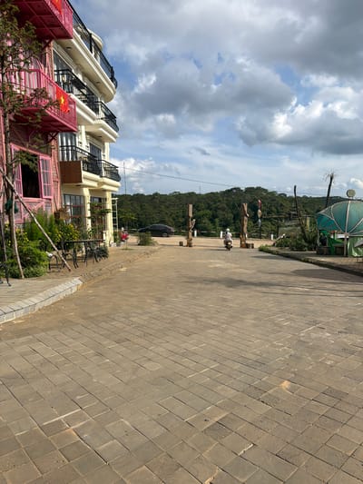 The image displays an outdoor street scene in Măng Đen, Vietnam, under a bright, partly cloudy sky. The perspective is taken from a wide, tiled pavement on the right, looking down a paved road that extends into the distance.

On the left side of the road, a row of multi-story buildings is visible. The lower sections of these buildings feature striking pink and red corrugated metal facades, adorned with several Vietnamese flags (red with a yellow star). Above, the buildings transition to cream-colored exteriors with elegant black, ornate balcony railings. In front of these structures, a small garden area bursts with green plants and bright yellow flowers, alongside outdoor tables and chairs, suggesting a cafe or restaurant.

Along the paved road, a person is seen riding a white scooter, heading away from the viewer. Further down, a black car and a red car are parked. The roadside is decorated with large, rustic wooden posts, some of which appear to have lanterns or other embellishments. In the mid-ground, a distinctive light green or teal-colored gazebo-like structure stands out.

The background is dominated by a vast expanse of densely forested hills, indicating a mountainous or hilly setting. The sky overhead is a mix of light blue patches and large, fluffy grey and white clouds. The lighting suggests it is daytime, casting clear shadows on the pavement. The scene combines urban or commercial development with a rich natural landscape. No readable text is visible other than the emblem on the Vietnamese flags.