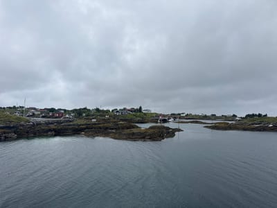 The image depicts a panoramic view of a coastal settlement on Sleneset, Norway, under an overcast sky. In the foreground, dark, rippling water stretches across the frame, leading towards a cluster of rocky islands dotted with sparse vegetation. These islands are intricately connected to the mainland, forming a natural harbor or bay. On the mainland and the islands, a small village of red and white houses is visible, nestled amongst the rocky terrain and green hills. Several boats are anchored in the calm waters, and a few small sailing vessels are present. The architecture of the houses suggests a traditional Scandinavian coastal style. The overall atmosphere is serene and maritime, indicative of a quiet fishing village or a small community by the sea. There are no people visible in the image.