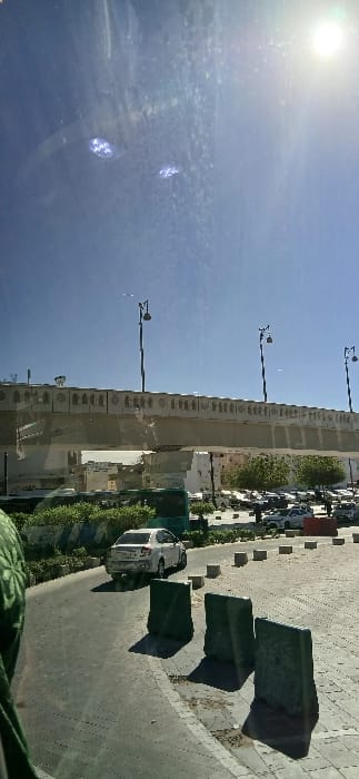 This image depicts a sunlit urban scene in the city of Medina, Saudi Arabia, viewed from inside a vehicle. The scene is dominated by a prominent, light-colored elevated concrete structure spanning horizontally across the mid-ground. This structure, likely an overpass or elevated roadway, features decorative panels along its side with repetitive arched cutouts. Mounted atop the elevated structure are at least three tall, dark street light poles, each fitted with double lamp fixtures.

Below the elevated structure, the ground level features a paved road curving from the lower left towards the center-right. A single white sedan is observed traveling on this road, moving away from the viewer. To the right of the road, a paved pedestrian area or plaza is delineated by a series of three dark green, rectangular concrete barriers, casting distinct shadows to their left. Beyond these barriers and stretching towards the background is a parking area filled with numerous parked cars, predominantly light-colored.

Green shrubs and trees are visible lining parts of the road and within the urban landscape. In the background, beneath and beyond the elevated structure, several multi-story buildings of varying heights and light coloration are visible. The sky above is clear and blue, with a bright sun flare visible in the upper right quadrant of the image, indicating strong sunlight and casting sharp shadows across the scene. A partial view of green fabric in the lower-left foreground suggests the photograph was taken from within an enclosed space, such as a vehicle.