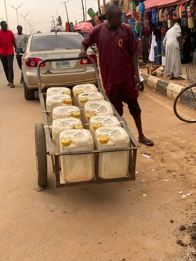 A street scene in what is likely Kafanchan B, Nigeria, displays a man pulling a hand-pulled cart loaded with twelve white plastic jerrycans along the road. The road is dusty and appears unpaved in sections. The man wears a maroon outfit and sandals. A beige sedan, with license plate NAS-480MB, is beside the cart. The rear of the car is visible. The background includes street vendors displaying goods under tents. Other people are walking nearby. Overhead power lines are visible.
