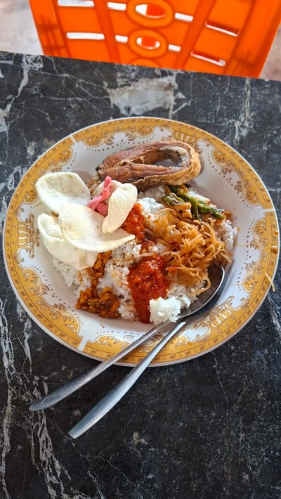 A plate of rice is served with fried fish, crackers, stir-fried vegetables, and sambal on a dark marble table. The plate has a decorative yellow and white pattern. Two spoons are placed on the table next to the plate. In the background, an orange plastic chair is visible. The scene appears to be set in Keudebayu, Indonesia, likely a local eatery.