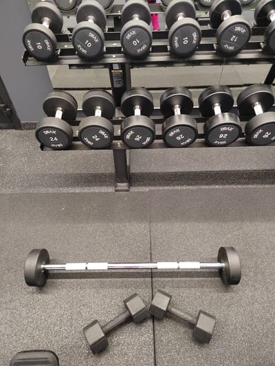 In a gym located in Novogorelovo, Russia, a variety of weightlifting equipment is displayed on racks and the floor. The upper rack holds dumbbells labeled with weights ranging from 10 to 12. The middle rack features dumbbells labeled 24 and 26. On the floor, a barbell with black end weights rests horizontally, accompanied by two smaller, black dumbbells with textured handles. The environment appears to be a clean, well-lit fitness facility, with dark grey rubber flooring and a dark grey wall in the background. There are no people visible in the image.