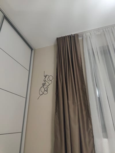 The image shows an interior scene. On the left is a white wardrobe with horizontal panels and silver trim. Adjacent to the wardrobe, on a beige wall, is a black, single-line art piece depicting two faces. To the right of the artwork are heavy, brown curtains next to semi-sheer white curtains. The location is likely in a room in Mosomishte, Bulgaria.