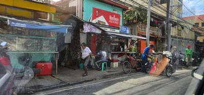 The image depicts a bustling street scene in Manila, Philippines, likely during the daytime given the bright sunlight casting shadows. The setting is a narrow urban street lined with small shops and residential buildings.

The primary subjects are several people and various objects common in a commercial area. On the left, a stall displays goods under a blue tarp with "G Gcash" visible. Next to it, a red cooler and a fan are present. Further down the street, a small store named "LINDA STORE" is visible with a prominent "Marlboro" advertisement. Several people are seen interacting around these establishments.

One man in a white shirt and gray pants walks along the sidewalk. Another person, wearing a black shirt with a white skull design, sits on a green stool. A man in a blue shirt is tending to a bicycle with a cargo trailer. Another man in a bright green shirt, holding a shovel, stands on the right side of the road, suggesting some sort of street work or maintenance is occurring.

Numerous motorcycles are parked along the street, indicating a common mode of transportation. The scene is characterized by dense overhead electrical wires, a common sight in the Philippines, adding to the urban and somewhat cluttered atmosphere. The overall impression is one of everyday life in a busy, working-class district, with commercial activity and daily routines unfolding on the street. The image conveys a sense of lively, unscripted street life.