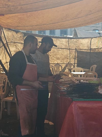 Two men stand under a tent. The man in front wears a pink apron over a black shirt. The other man wears a gray shirt and a black baseball cap. Tables and chairs are visible in the background, and a building stands in the distance. An orange canopy obscures some of the scene.