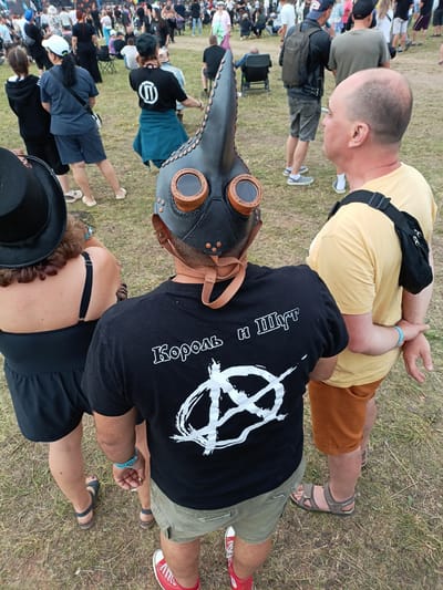 The image shows a group of people at an outdoor event, possibly a music festival, in Lagunovo, Russia. In the foreground, a person wearing a black plague doctor mask with goggles is standing and facing away from the camera. They are wearing a black t-shirt with a white graphic design and Cyrillic text. To the left of this individual is a person wearing a black hat and a black dress. To the right, a man wearing a yellow t-shirt and orange shorts is standing with his arm outstretched, seemingly interacting with the person in the plague doctor mask. Behind these foreground subjects, a crowd of people is visible, some sitting and others standing, with tents and staging in the background. The environment appears to be a grassy field.
