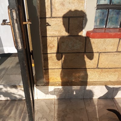 This image, originating from Mahiakalo ward, Kenya, displays an exterior residential or commercial scene under direct sunlight. A dark-colored door frame is visible on the left, with an open door featuring a brass-toned handle and lock mechanism. The central and right portions of the image feature a building wall constructed from light brown or tan rectangular blocks with dark mortar. A prominent human-shaped shadow is cast upon this wall and the tiled ground, depicting an individual with a distinctive head outline and arms slightly bent. To the upper right, a window with dark frames and multi-paned glass, exhibiting a blue tint in some sections, is visible above a red-colored sill. The ground is covered with light-colored, mottled grey and white patterned tiles. In the lower right, a distinct animal-like shadow, resembling a dog's head and ear, is cast on the tiles, with a dark, furry tail visible at the extreme bottom right.