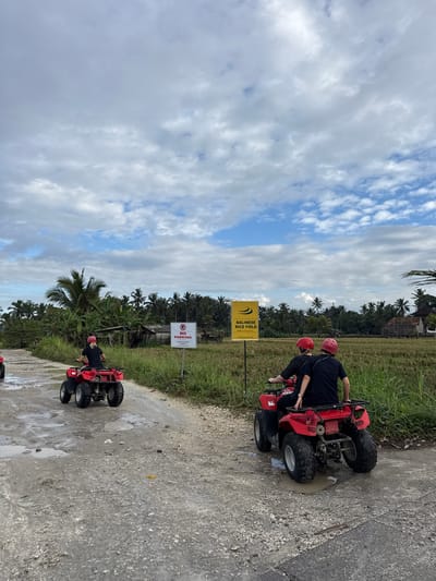 In Payangan, Indonesia, two people are riding a red ATV on a muddy dirt road. They are wearing red helmets. The person in front is steering, and the person behind is holding onto the driver. In the background, there is a field of rice paddies, palm trees, and a few buildings. A yellow sign with the words "BALINESE RICE FIELD" is visible on the left side of the road. Another sign with "NO PARKING" is on the right side. The sky is cloudy with patches of blue.