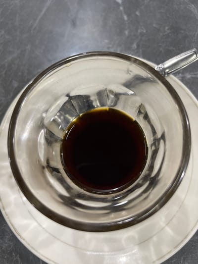 The image is a top-down view of a clear glass cup filled with dark liquid, likely coffee, sitting on a white saucer. The saucer is placed on a gray, marbled surface. A metallic spoon is partially visible to the left of the cup, resting on the saucer. The lighting suggests an indoor setting, possibly a cafe or dining area, with reflections visible on the glass of the cup. There are no people or text visible in the image. The scene is static, with no activity taking place. The overall impression is one of a simple, focused composition on a drink.