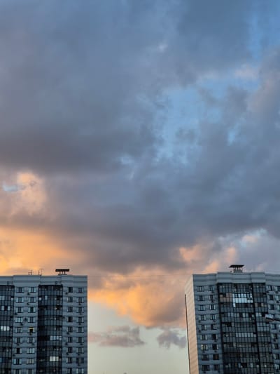 The image shows two modern apartment buildings against a dramatic sky with an approaching storm. The buildings are tall, with many windows and balconies, and appear to be identical in design. The sky is filled with dark, ominous clouds, with streaks of orange and pink light breaking through from the setting sun. A thin wire stretches horizontally between the two buildings. The scene suggests an urban environment in Voronezh, Russia, likely during the late afternoon or early evening. There are no people visible in the image.