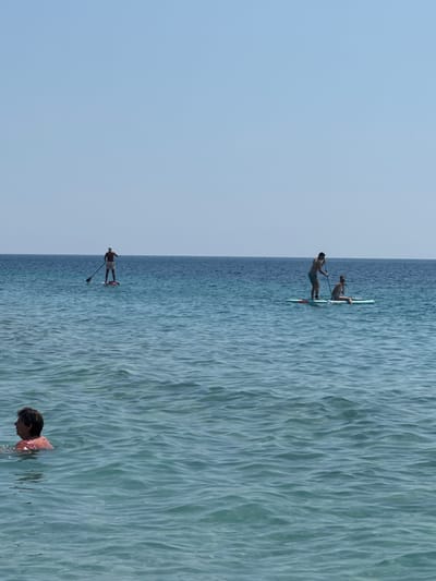 The image shows a serene scene at Mirište, Montenegro, with calm, rippling turquoise waters under a clear blue sky. Several people are engaged in various water activities. In the foreground, a person is partially submerged in the water, their head and shoulders visible, wearing a pink life vest. Further out, two people are on a paddleboard together, one kneeling and the other standing, both appearing to be enjoying a leisurely paddle. Even further in the distance, another individual is standing on a paddleboard, holding a paddle. The overall atmosphere is peaceful and recreational, suggesting a pleasant day at the beach or a calm body of water. The time of day appears to be daytime, with ample sunlight illuminating the scene, and the weather seems to be fair and warm, ideal for these outdoor activities. The clear sky and gentle waves contribute to the tranquil ambiance.