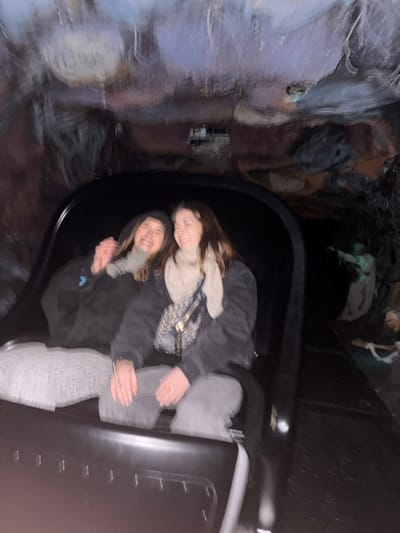 Two adult females are seated in a dark-colored ride vehicle, likely within Phantom Manor at Disneyland Paris in Chessy, France. Both individuals are laughing, their mouths open. The female on the left wears a dark hooded garment and a light-colored scarf, with her left arm partially raised. The female on the right wears a dark jacket, a light-colored scarf or turtleneck, and light gray pants, carrying a patterned cross-body bag. The background consists of a dimly lit, artificial cavernous environment with blurred rock formations on the walls and ceiling. To the right of the subjects, an illuminated, pale animatronic figure resembling a ghost or zombie with outstretched arms is visible, along with a partially obscured skeletal figure further right. The image exhibits motion blur, consistent with the vehicle being in motion.