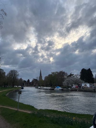 The image shows a scene in the Vale of White Horse, United Kingdom. A river flows from bottom to top, with vegetation and a path on the right bank, and buildings and boats along the left bank. The sky is predominantly filled with grey and white clouds. In the middle ground, a church spire is visible among the buildings along the left bank. The foreground shows vegetation at the bottom. A metal post is on the right bank. The scene appears to be taken on an overcast day.