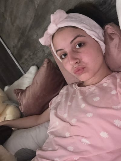 The media file shows a close-up, rotated view of a person lying down. The subject is wearing a pink t-shirt with a delicate white daisy pattern and a light pink headband with a bow. Their face is turned towards the camera, with their lips pursed in a playful expression. The person appears to be in a relaxed or comfortable setting, as suggested by the pillows around them, including a dusty rose velvet pillow and a white pillow. The background is a dark, textured surface, likely a floor or furniture. The lighting suggests it could be indoors during the daytime, though the exact time is not discernible. No visible text is present in the image. The overall mood is casual and intimate, possibly indicating a moment of downtime or self-care.