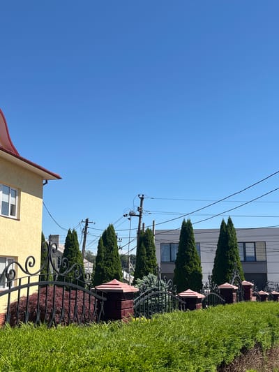 This is a vertical, eye-level shot of a residential area on a sunny day in Chahor, Ukraine. The bright blue sky dominates the left half of the image, with power lines stretching across the top of the frame. To the right, a neat green hedge runs along a black wrought-iron fence, with decorative brick pillars topped with pointed red caps. Behind the hedge and fence, several tall, cone-shaped evergreen trees are spaced out. Beyond the trees, a modern, grey, multi-story building with large windows is visible. In the foreground on the bottom right, the corner of a yellow building with a red-tiled roof and a white-framed window is visible. The lighting suggests it is daytime, likely morning or afternoon given the bright sun. There are no people visible in the frame, and the scene depicts a tranquil, well-maintained urban or suburban environment.