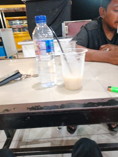 The image depicts a casual scene at an outdoor eatery in Cotpuuk, Indonesia, likely in the evening or at night, given the artificial lighting visible. The primary focus is a table with several items on it. A person, partially visible on the right side of the frame, is wearing a dark t-shirt and appears to be a man. He has a ring on his right hand. In front of him are two clear plastic cups, one with a light-colored beverage and a straw, and another appearing empty or mostly empty. A plastic bottle of water with a blue cap stands upright. Also on the table are what appear to be keys and a small object, possibly a wallet or coin purse, next to a stapler. A bright green lighter is visible on the right side of the table.
In the background, there's a glimpse of what could be a food stall or cart with yellow and white structures. The overall atmosphere suggests a relaxed, possibly social gathering. The lighting is dim, and the background is out of focus, emphasizing the objects on the table. There is no visible text in the image that can be clearly deciphered.
