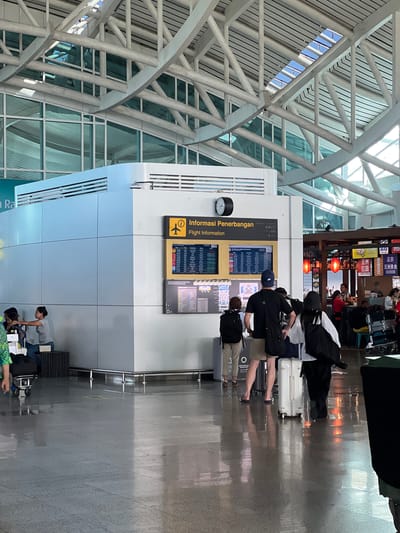 This is an image taken inside an airport terminal in Kuta, Indonesia, characterized by its modern architectural design with exposed white beams and a high ceiling featuring a skylight. In the mid-ground, a white structure houses flight information displays. Two screens are visible; the top one, labeled "Informasi Penerbangan" (Flight Information), shows a list of flights with details such as flight numbers, destinations, and times. Below it, another screen displays a map of the terminal. Several people are present in the scene. In the foreground, a group of individuals is walking away from the camera, some with luggage. To the left, a woman is seated and looking to her right. Further back, a counter with food establishments is visible, with staff attending to customers. The floor is polished and reflects the light, adding to the bright and open atmosphere of the terminal.