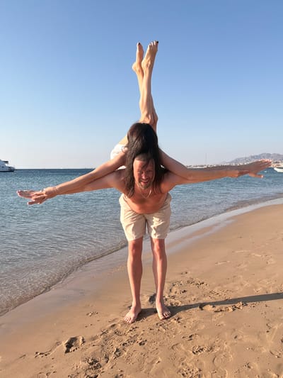 An outdoor, full-shot photograph captures two individuals performing an acrobatic or yoga-like pose on a sunny beach in Safaga, Egypt. The image appears to be taken during the golden hour, with warm light illuminating the scene and casting long shadows on the sand.

The primary subject, an adult with long dark hair and a beard, serves as the base for the pose, standing on the sandy shore with their legs spread horizontally. They wear a light-colored top and khaki shorts and are smiling directly at the camera. Their arms are also extended horizontally, supporting another person, whose bare legs are visible extending horizontally to the left from the primary subject's torso. The second person's feet have dark-painted toenails.

The picturesque setting features a calm, blue sea with gentle waves lapping the sandy beach. In the background, majestic mountains and coastal buildings are visible, with boats dotting the distant horizon on the water under a clear blue sky. The sandy beach shows various footprints and ripple patterns from the receding tide. The overall atmosphere conveys joy and athleticism. No text is visible in the image.