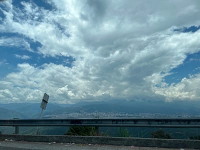 The image displays an elevated, panoramic view of the city of Girón, Colombia, observed under a partly cloudy sky. In the immediate foreground, a concrete curb or barrier extends horizontally across the bottom edge, showing evidence of scattered debris. Directly behind this, a silver-colored metal guardrail spans the width of the frame. Positioned between the guardrail and the distant cityscape, a rectangular metal sign, light in color and appearing weathered, is significantly tilted to the left on its pole. Sparse green vegetation is visible behind the guardrail. The midground encompasses the extensive urban sprawl of Girón, characterized by a dense concentration of buildings that stretch across the horizon. Beyond the city, multiple layers of mountains with blue-green hues recede into the distance, partially obscured by atmospheric haze. The upper two-thirds of the frame are occupied by a sky featuring numerous large, predominantly white and light-grey cumulus clouds, with distinct patches of clear blue sky visible in the upper left and right areas. No individuals, specific actions, or interactions are discernible.