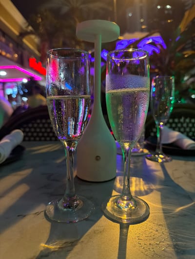 Two champagne flutes are prominently displayed on a marble-topped table, one in the foreground and another slightly behind and to the right. Both glasses contain a light-colored liquid, with condensation visible on the exterior of the left flute. The right flute has a foamy layer at the top. In the background, a white cylindrical object with a circular top is situated between the glasses. The scene is illuminated by colorful, out-of-focus lights, including pink and purple neon, suggesting an urban nightlife setting. Shadows cast by the glasses and the central object stretch across the table. The blurry backdrop features palm fronds and architectural elements, consistent with a city environment. The overall ambiance is one of an evening outing, likely at a bar or restaurant in Sunny Isles Beach, Florida, United States of America.