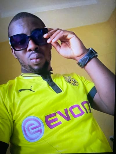 A man wearing a yellow Borussia Dortmund soccer jersey and dark sunglasses is photographed close-up. He has a watch on his left wrist. The jersey displays a purple "EVONIK" logo. He is holding the sunglasses with his left hand. The background is plain and light-colored. The image is slightly rotated.
