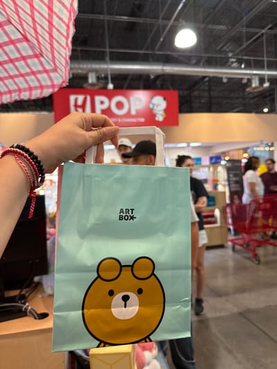 The image shows a person holding a light blue "ART BOX" shopping bag with a bear cartoon on it. The person is wearing multiple bracelets. The background includes a store setting with shelves and people, some with shopping carts. A red sign with "POP" and a cartoon character is also visible. The location is Orlando, United States, based on the ART BOX store presence in that city.