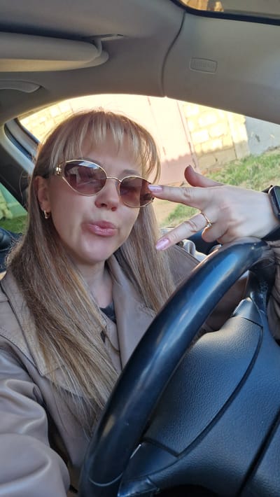 A fair-skinned woman with blonde hair is in the driver's seat of a car, wearing sunglasses and a beige leather jacket. Her left hand is raised with a ring on her finger. The steering wheel is visible, and an "AIRBAG" label is on the car's interior. The location is Otchor, Russia. The photograph appears to be taken from inside the vehicle.