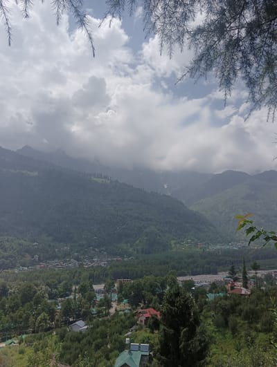 The image captures a panoramic view of a valley with lush green hills and mountains under a cloudy sky. In the foreground, branches of pine trees hang from the top, framing the scene. A river or stream can be seen flowing through the valley, with a town or village nestled amongst the trees. Buildings with red and grey roofs are visible, as are some larger structures and a few roads. The distant mountains are shrouded in mist and clouds, with patches of snow visible on their upper slopes. The overall impression is one of a serene and picturesque natural landscape combined with a human settlement. The location is identified as Bahang, India, suggesting a mountainous region in the Himalayas.