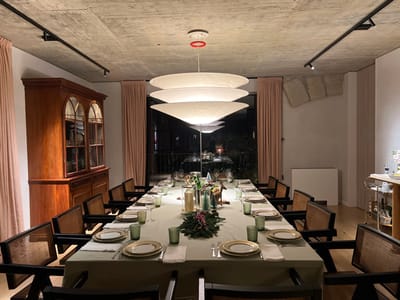 An indoor shot depicts a formal dining area with a long table set for a meal. The table features a light green tablecloth, plates, glassware, and silverware. A centerpiece adds a decorative touch. Surrounding the table are chairs with dark frames and woven backs. The room includes a wooden cabinet, curtains, track lighting, and a unique, layered chandelier. A window reveals a glimpse of the outdoors at night. A bar cart is visible in the background.