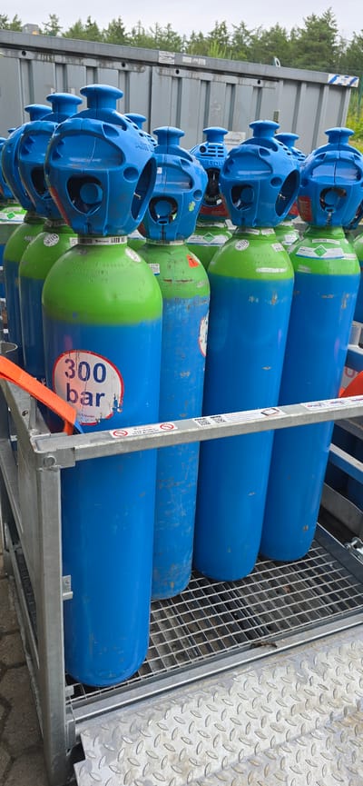 In Haren (Ems), Germany, a metallic cage-like structure holds a vertical arrangement of gas cylinders. The cylinders are predominantly blue with green tops. One prominent cylinder in the foreground displays a white circular label with "300 bar" printed in bold blue letters. The cylinders appear to be stored outdoors, with a grey industrial-style building and green trees visible in the background. An orange strap is attached to the cage structure, hinting at a loading or securing mechanism.