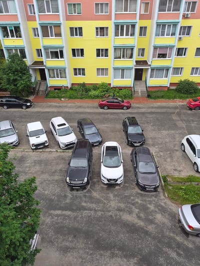 This is an aerial, top-down view of a parking lot next to a brightly colored apartment building in Nyagan, Russia. The building is painted in sections of yellow, pink, and white with blue trim around the windows. Many cars are parked in the asphalt lot, including a red sedan, several dark-colored sedans and SUVs, and a few white vehicles.
The scene appears to be in the daytime, with natural light illuminating the area. There are no people visible, but the presence of parked cars suggests it is a residential area. Green foliage and trees surround the parking lot and are visible along the side of the building. The asphalt appears damp, indicating recent rain or wet conditions.
Visible text includes license plates on some of the vehicles, but they are not legible enough to be transcribed. The overall impression is a typical urban or suburban parking area.