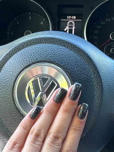 The image displays a close-up view of a person's hand with glittery black nail polish resting on the steering wheel of a Volkswagen car. The car's dashboard is visible, showing a speedometer with markings up to 140, and a digital display indicating "17:19" along with a car icon with an open door. The steering wheel has the Volkswagen logo in the center. The background is out of focus, suggesting an indoor setting, potentially a car showroom or a private garage. There is no indication of the specific location within Razgrad, Bulgaria, or any identifiable landmarks.