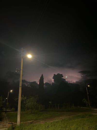 At night in Lozarevo, Bulgaria, a vertical shot shows a lightning storm behind a tree line. A concrete utility pole with a glowing light is in the foreground, illuminating grass and a narrow road. Power lines extend from the pole into the darkness.