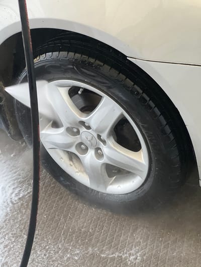 In the city of Shchelkino, Ukraine, a white car is being washed. The image focuses on the front passenger-side wheel and tire. A black hose, likely connected to a pressure washer, is visible in the foreground, spraying water onto the tire and rim. Water is actively splashing around the wheel, indicating the washing process is underway. The car's body, painted in a bright white, is partially visible above the wheel arch. The ground beneath the car appears to be a textured concrete surface, wet from the washing.