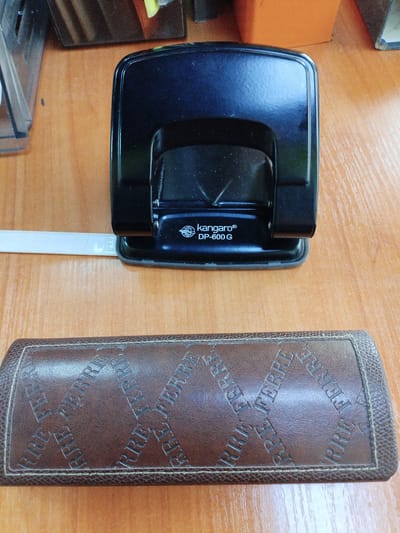 The image depicts a close-up, overhead view of a wooden desk surface. Prominently featured is a black Kangaro DP-600 G hole punch positioned in the upper-center of the frame. To the left of the hole punch, a white strip of paper is partially visible. Below the hole punch, a brown leather case with embossed lettering, appearing to be "FERRE" repeated in a diamond pattern, occupies the lower portion of the image. The background shows a blurred orange object and a portion of a glass object, suggesting an office or study environment. The location is identified as Вароніцкі сельскі Савет, Belarus, though the image itself provides no direct visual cues to confirm this specific locale.