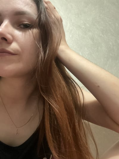 This media file is a close-up, low-angle photograph of a person's face and upper body, oriented sideways. The subject appears to be a young woman with long, reddish-brown hair that falls across her face and chest. Her eyes are open and looking slightly to the left, and her lips are parted. A delicate necklace with a small, geometric pendant is visible on her collarbone. Her left arm is bent and resting on her head, with her hand near her temple, suggesting a relaxed or pensive pose. The background is a textured, light-colored surface, possibly a wall or fabric, with a subtle marbled pattern.

The lighting suggests an indoor setting, likely during the day, given the soft, diffused light. There are no obvious external environmental cues like weather or specific landmarks that would pinpoint the location beyond the provided context of Kirov, Russia. The overall mood is intimate and personal, with a focus on the subject's facial features and hair. There is no visible text in the image. The cropped images highlight details such as a small mole on the subject's arm, the texture of the background, and the close-up of the subject's eye and necklace.