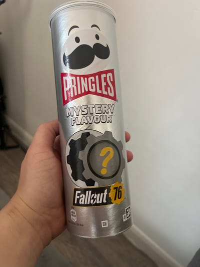 A hand holds a silver Pringles can. The can is labeled "Pringles Mystery Flavour" with a "Fallout 76" logo that includes a yellow question mark inside a gear. Nutritional information is printed near the bottom of the can. The background is a plain white wall. The image does not contain any information about Gorleston-on-Sea, United Kingdom.