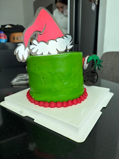 A green cake with red trim sits in a white box. It is decorated with a wooden Grinch-themed cake topper. One side shows the Grinch's red hat and white fur trim, while the other features his green hand holding a bag. The cake and box are on a black surface, likely a countertop. The image has been rotated to be portrait.