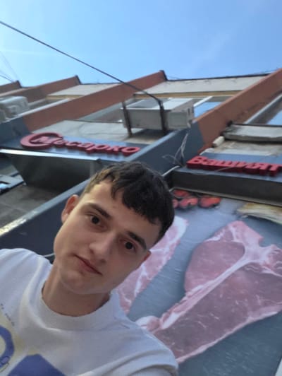 An outdoor, eye-level, medium shot shows a young Caucasian male with short, dark hair, dark eyes, and fair skin, looking towards the camera. He is wearing a white t-shirt with a graphic design on the left side of the chest. The background features the facade of a building with red lettering that spells "Багрят" (Bagryat) on a blue sign, and another sign with red lettering that spells "Четири" (Chetyre) above it. Below the signs, there is a large graphic advertisement of raw meat, specifically T-bone steaks, on a blue background. The building's architecture includes orange and cream-colored diagonal beams and light-colored walls. The sky is clear and blue. The image was taken in the city of Voluyak, Bulgaria, likely in front of a butcher shop or meat market.