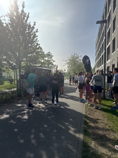The image shows a group of people gathered on a paved path next to a building in what appears to be an outdoor event or gathering in Antwerp, Belgium. A large New Balance branded flag is situated among the group. Some individuals are standing, while others appear to be seated in a wheelchair. There is a building with a grid-like window pattern in the background, along with trees and grassy areas. The sunlight casts shadows on the path.