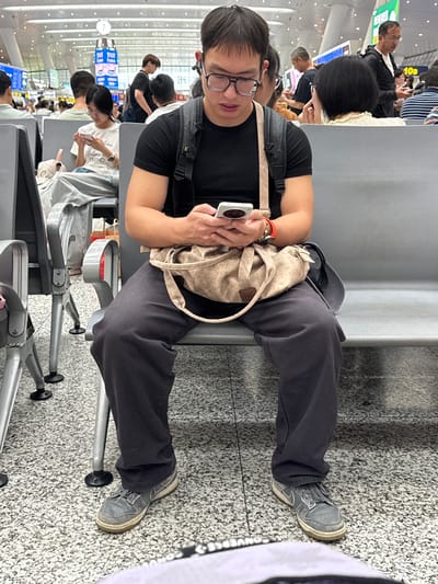 The image depicts a man sitting on a gray bench in a public waiting area, likely an airport or train station, given the overhead lights, signage, and seating arrangement. He is wearing a black t-shirt, dark gray sweatpants, and gray sneakers. He has glasses on and is looking down at a white smartphone in his hands. A beige shoulder bag is draped across his lap, and a black backpack is visible on his back. Other people are seated in the background, some looking at their phones. The floor is a speckled gray and white pattern. The environment suggests a bustling indoor public transit hub in Shangcheng District, China.