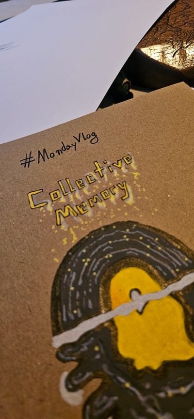 The image shows a close-up of a piece of brown cardboard with text and a drawing. The text "#MondayVlog" is written in black marker. Below this, "Collective Memory" is written in yellow marker with a black outline, with some yellow splatters around it. To the right of the text, there is a drawing of a broken vinyl record with a yellow center and black grooves speckled with yellow dots. The bottom portion of the drawing shows a dark, amorphous shape. The background is out of focus, showing a white paper on the left and a wooden surface with visible grain on the top right. There is no information in the image that suggests a location in Kostenets, Bulgaria.