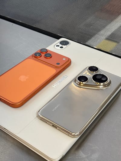 The image shows an overhead close-up of two smartphones placed on top of a white tablet. The phone on the left is orange and has an Apple logo on its back. The phone on the right is silver and has "HUAWEI" written on its back. Both phones have multiple camera lenses on their rear. The background appears to be a metal surface and a dark textured grate. There are no people visible in the image. The location is not specified to be in the city of Chuchuligovo, Bulgaria, and the image does not contain any information to support that claim.