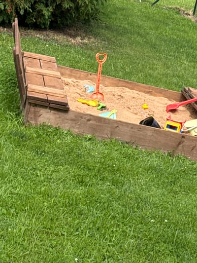 The image shows a wooden sandbox filled with sand and children's toys, set in a grassy yard. The sandbox is rectangular with wooden plank sides and a built-in wooden bench on one side. Inside the sandbox, there are several toys including a red shovel, a yellow duck, a blue bucket, a yellow sand mold, and an orange toy rake. A toy truck is also visible. The sand is piled up in some areas. The yard is covered in green grass, with some small white flowers scattered throughout. To the left of the sandbox, there is a dark green shrub. The scene appears to be outdoors on a sunny day, judging by the lighting and the vibrant green of the grass. The overall impression is one of a playful outdoor setting, likely a backyard in a residential area. The location context provided is Kārsava, Latvia. There is no visible text in the image. The image is taken from a high angle, looking down into the sandbox and the surrounding yard.