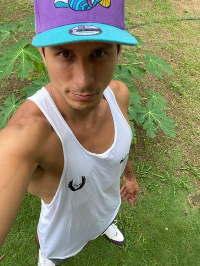 This is a high-angle, vertical, selfie-style photograph of a person wearing a purple and teal baseball cap and a white tank top. The person is looking directly at the camera, with a neutral expression. Their left arm is extended, holding the camera. Their right arm is bent, resting on their side. Visible text on the baseball cap reads "NEW ERA SNAPBACK". The person is standing outdoors on a grassy area with large green leaves from a plant visible in the foreground. The lighting suggests it is daytime, likely outdoors in a tropical or subtropical environment. The background consists of more grass and some dirt. The person's skin tone is medium. The overall scene is casual and appears to be taken in a park or garden. The individual has a tattoo on their left forearm. The person is wearing white and red athletic shoes.