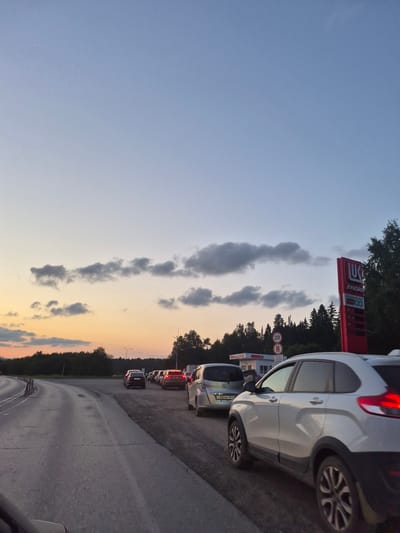 The image shows a queue of cars on a road near a Lukoil gas station. The sky is a gradient of light blue and orange, with scattered clouds. The gas station sign is red with white lettering and a yellow logo. There are trees and a forest in the background. The road is paved and appears to be a two-lane highway. There are no people clearly visible in the image. The scene appears to be at dusk or dawn.