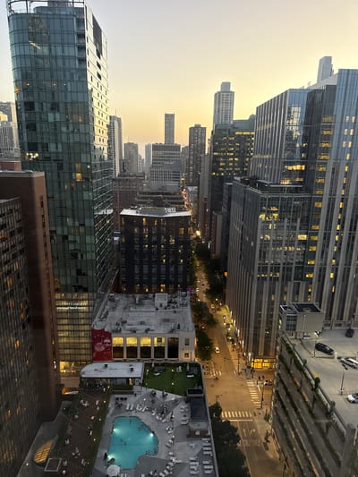 This is an aerial, eye-level, vertical, full shot of a city skyline in Chicago, Illinois, United States of America, taken at dusk. The sky is a gradient of pale orange and yellow, indicating the sun is setting. The foreground is dominated by the side of a tall, modern building with a dark, brick-like facade. To the right, a large, curved glass building reflects the twilight sky and surrounding city lights.
Below these buildings, a street is visible with traffic, streetlights, and pedestrian crosswalks. There are several other tall buildings of varying architectural styles, some with illuminated windows, extending into the distance. A multi-level parking garage with cars parked on its levels is on the right side of the image.
Further down, a rooftop amenity space is visible. This area includes a swimming pool with people swimming, lounge chairs around the pool, a grassy area with seating, and a walkway lined with string lights. People are seen scattered around this outdoor space. A large red mural of a man's face is visible on the side of one of the buildings. The overall scene depicts a bustling urban environment as day transitions into night.