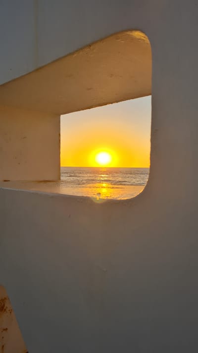 A low-angle shot from within a building shows a rectangular opening with rounded corners, framing a view of the sunset over the sea. The interior walls are painted a light off-white color, and the opening is slightly discolored by age or wear. The sun is bright yellow, positioned just above the horizon, casting a shimmering reflection on the water. This is likely a scene from Nahariyya, Israel, given the image title. There are no people visible in the frame.