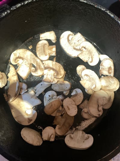 An overhead, close-up shot captures a collection of sliced mushrooms being cooked or prepared in a dark, round pan, likely a cast iron skillet. The pan's surface is black, textured, and shows some areas of dried white residue, possibly from previous cooking or cleaning.
The primary subjects are numerous white button mushrooms (Agaricus bisporus), cut into various slices and pieces, scattered across the bottom of the pan. Some slices are oval, while others are more irregular, revealing the internal structure and gills. A few mushrooms exhibit a slightly browned or translucent appearance, suggesting they are in the process of being cooked, possibly sautéed, with some liquid or oil visible reflecting light. Dark specks, potentially seasoning like pepper, are visible on some mushroom surfaces.
The lighting appears to be bright, casting some reflections on the wet surfaces of the mushrooms and the pan. There are no people visible, and no discernible text is present in the image. The scene is consistent with food preparation in a kitchen environment. While the location context provided is Novoe Devyatkino, Russia, there are no specific visual cues within the image itself to confirm this geographical location. The focus is entirely on the culinary process.